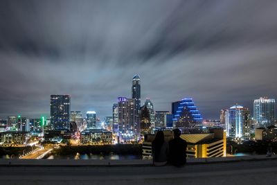 Man and woman looking at illuminated cityscape while sitting on terrace at night