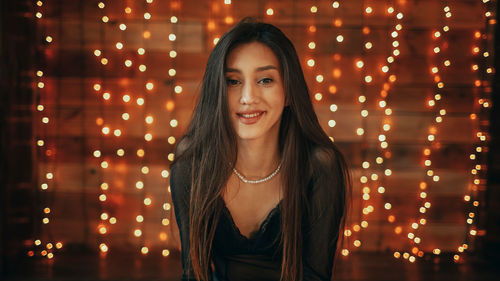 Portrait of young woman standing against illuminated string lights at night