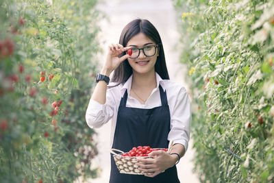 Portrait of smiling young woman holding fruits while standing amidst plants on field