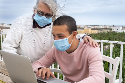 Boy wearing mask with grandmother using laptop outdoors