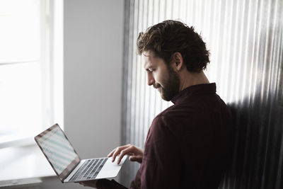 Businessman standing by corrugated iron wall while using laptop at creative office