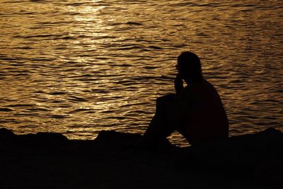 Silhouette woman sitting on rock by sea during sunset