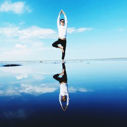 Full length of young woman meditating while standing on shore at beach against sky during sunny day