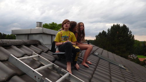 Siblings sitting on roof against cloudy sky during sunset