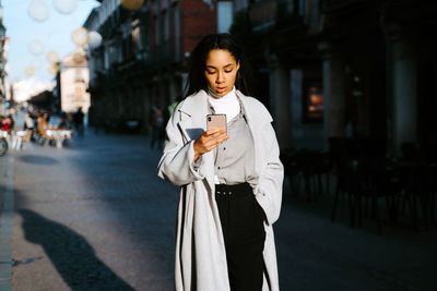 Young ethnic female in stylish casual outfit messaging on mobile phone while standing on urban street in sunny autumn day