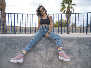 Determined female millennial in street style wear and with tattoos sitting on ground in skate park and looking at camera while relaxing at weekend