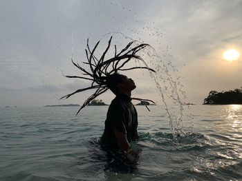 Man tossing dreadlocks in sea against sky during sunset