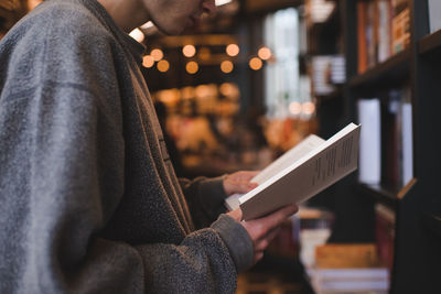 Teenager boy holding paper book standing in public library over bookshelf and glowing light