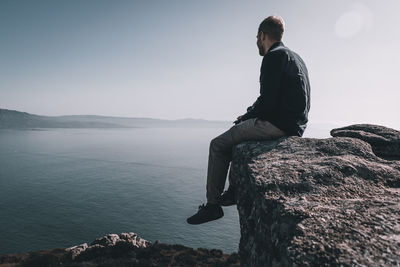 Man sitting on rock looking at sea and mountain against sky