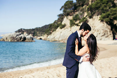 Newlywed couple kissing at beach against clear sky during sunny day