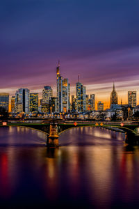 Illuminated bridge over river by buildings against sky at sunset