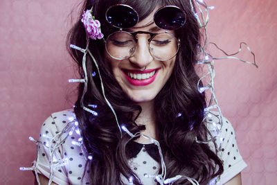 Close-up of smiling female model with illuminated string lights against peach backdrop