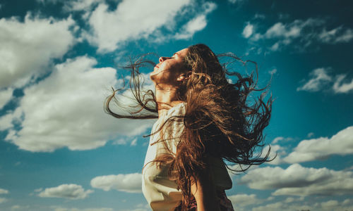 Low angle view of young woman with tousled hair standing against cloudy sky