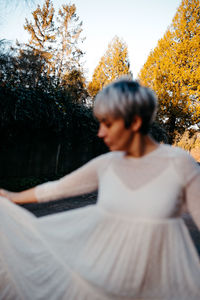 Side view of blurred anonymous female with short hair wearing white dress standing against tall green trees in rural terrain