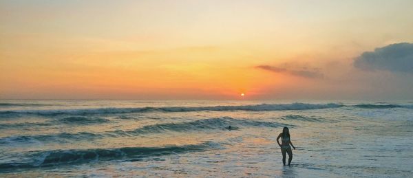 Woman wearing bikini while standing at beach against sky during sunset