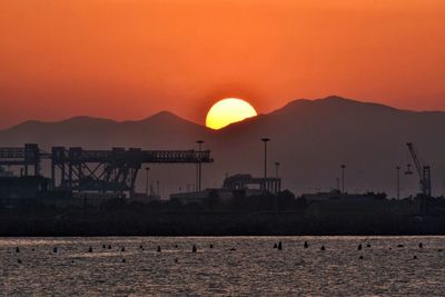 Silhouette factory by sea against orange sky