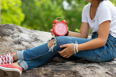Low section of woman holding alarm clock while sitting outdoors