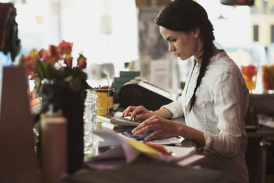 Side view of serious owner using calculator while looking at digital tablet in counter