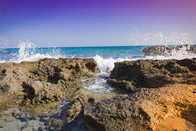 Waves splashing on rocks at shore against clear blue sky