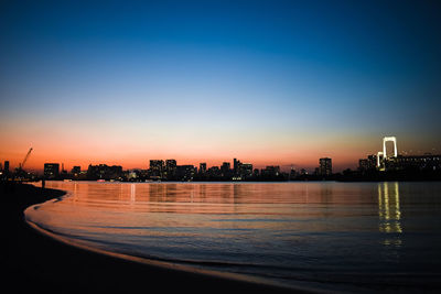 Illuminated buildings by river against sky during sunset