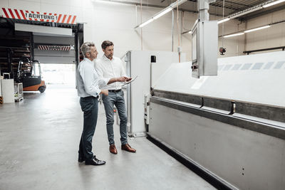 Male engineers discussing over digital tablet while standing by manufacturing equipment in industry