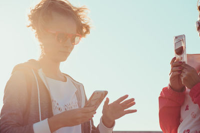 Low angle view of mother with daughter using mobile phones while standing against sky during sunny day