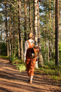 Woman botanist in orange overalls with backpack on ecological hiking trail in forest. 