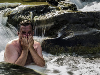 Shirtless mature man covering mouth in waterfall