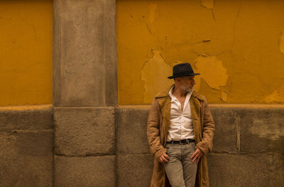 Portrait of adult man in hat and coat against yellow wall on street