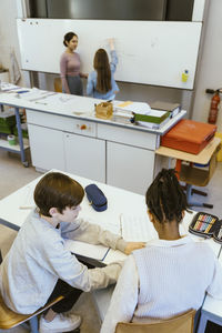 High angle view of boy helping male friend while sitting on desk in classroom