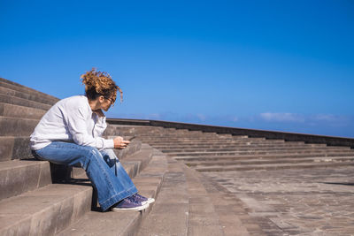Mature woman using mobile phone while sitting on steps against blue sky