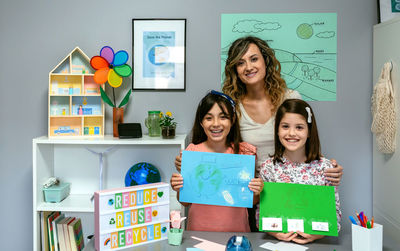 Teacher in classroom looking at camera next to students showing colorful ecological drawings