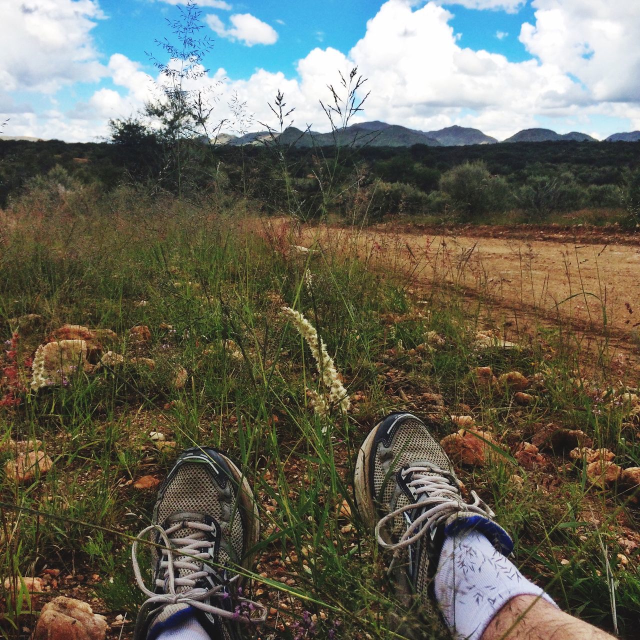 low section, grass, sky, person, field, landscape, personal perspective, one person, shoe, tranquility, tranquil scene, nature, cloud - sky, grassy, mountain, scenics, beauty in nature, cloud, part of