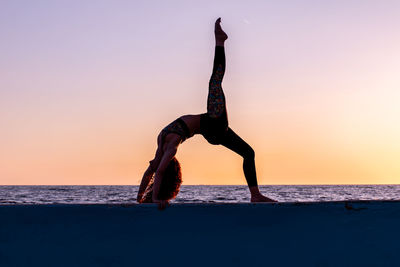 Full length of woman practicing yoga by sea against sky during sunset