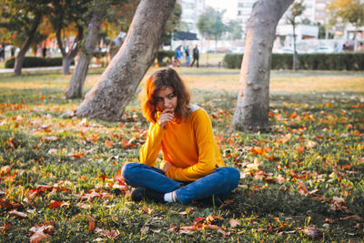 Curly young girl in yellow sweater and jeans sitting on fall grass with dry leaves.