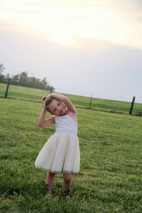 Portrait of cute girl with head cocked standing on field against sky during sunset