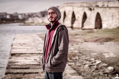 Portrait of young man standing against bridge over river