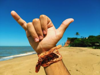 Close-up of man showing shaka sign at beach against blue sky