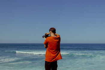 REAR VIEW OF PHOTOGRAPHER PHOTOGRAPHING SEA AGAINST SKY