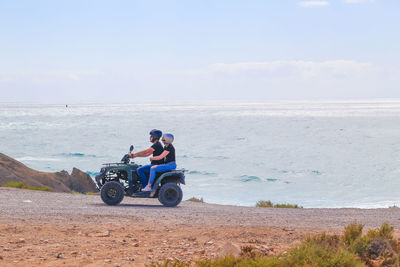 Side view of couple riding quadbike at beach against sky