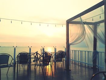 Empty chairs and tables by sea against sky during sunset