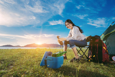 Portrait of smiling young woman camping on grassy field against blue sky