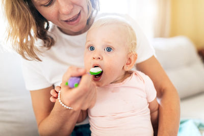 Crop smiling woman holding cute blond haired toddler and feeding with spoon in bright room on blurred background