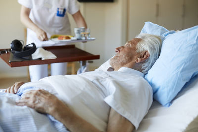 Senior man looking at female nurse serving breakfast while lying on hospital bed