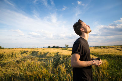 Side view of young man with eyes closed while standing on wheat field against sky