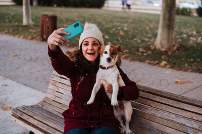 Woman with dog using phone while sitting on bench against trees