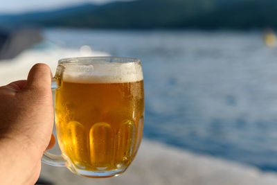 Hand holding mug of cold beer by sea