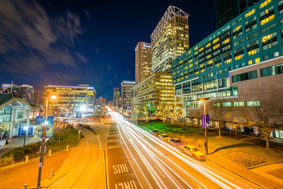 Light trails on road amidst buildings against sky at night