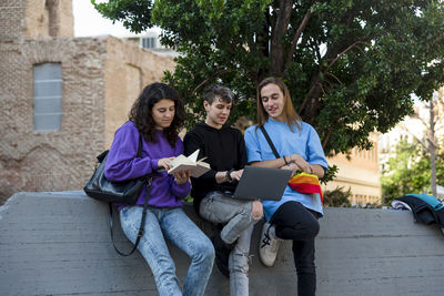 Young diverse people with lgbt rainbow flag using laptop and book outdoors.