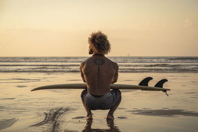 Young man with surfboard crouching at seashore during sunset